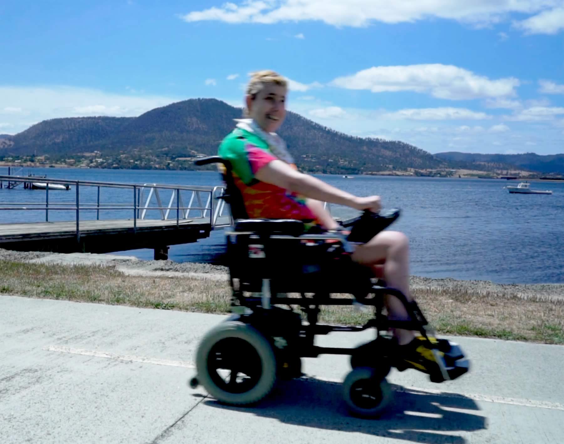 Still photo of Janelle McMillan in an electric wheelchair travelling along a footpath with the Derwent River in the background. Still taken from the video made by Lara van Raay for the Rafting project.
