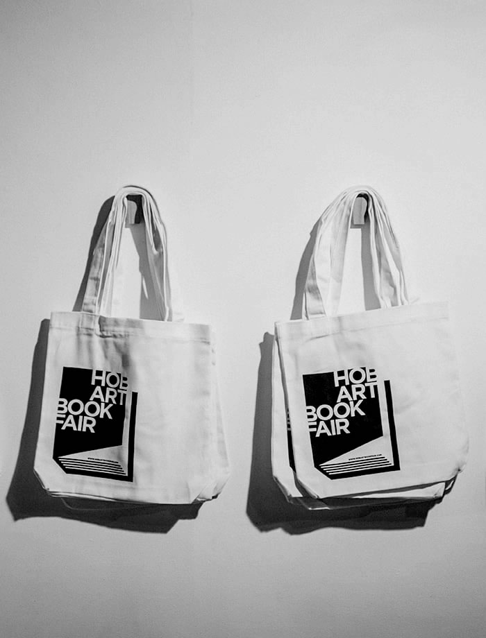 Black and white photo of bundles of white tote bags hanging from two hooks, with the Hob/ART book fair logo on them. Photo: Rosie Hastie for The People’s Library 2018.