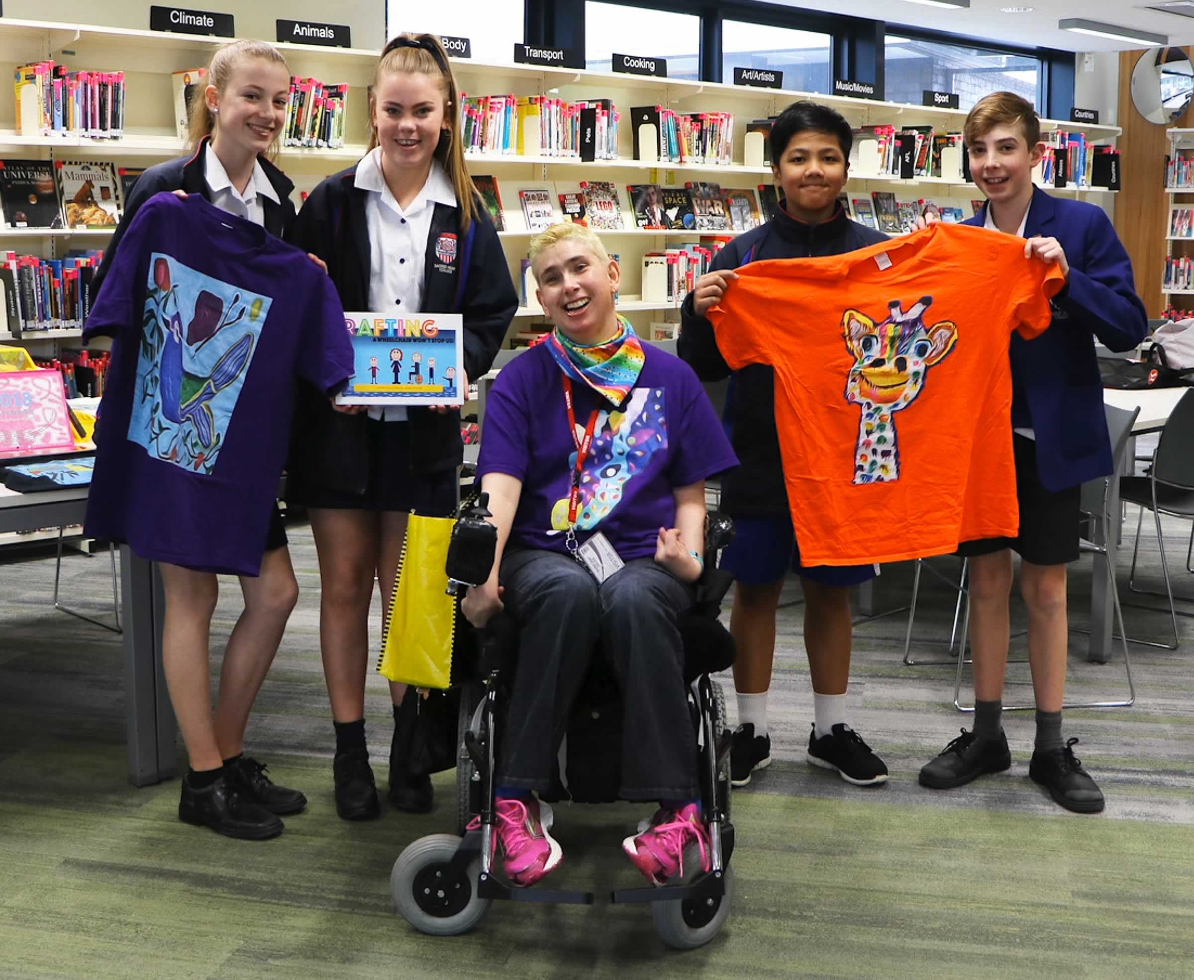 Photo of Janelle McMillan with four school children holding some of her t-shirts and books, with bookshelves behind them — all smiling.