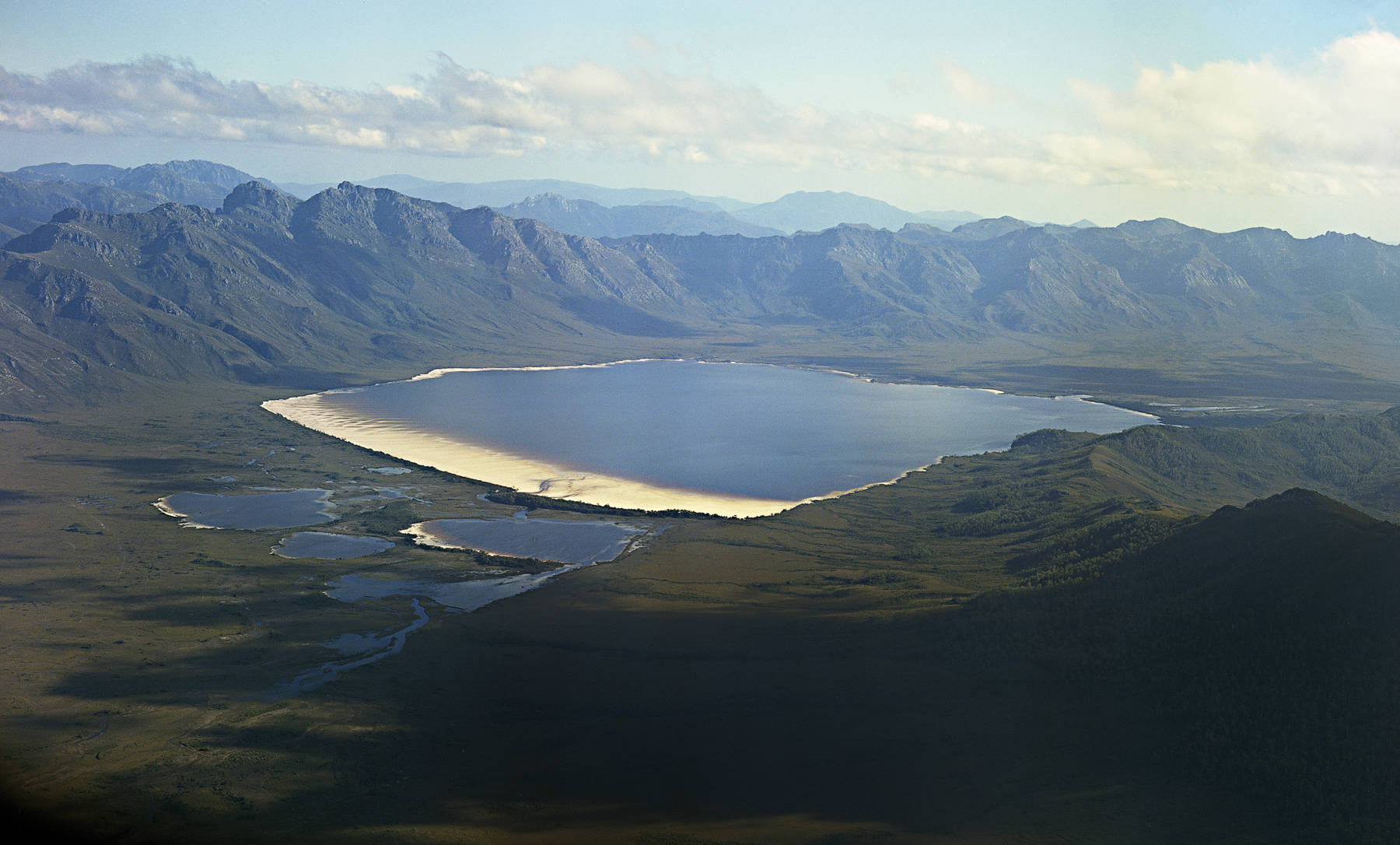 Photograph of the original Lake Pedder by Wilf Elvy taken from a light plane in 1972–73 prior to inundation by the Huon-Serpentine impoundment waters. The rugged Frankland Range is in the background and the three kilometre long, stunning pink quartzite beach sparkles in the foreground.