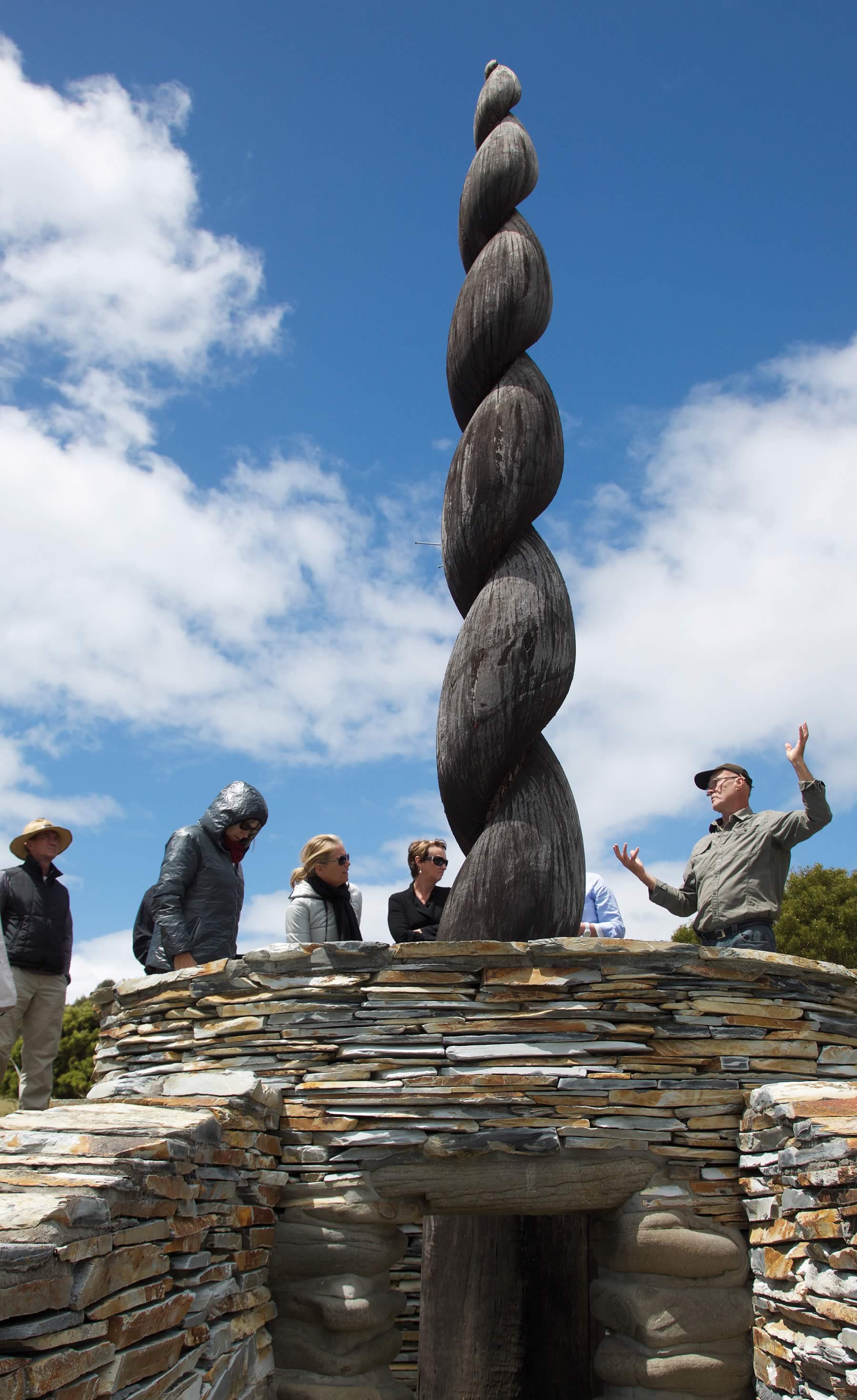 Photo of Peter Adams talking to a group of people standing around a stone and wood sculpture. Photo courtesy of Peter Adams.